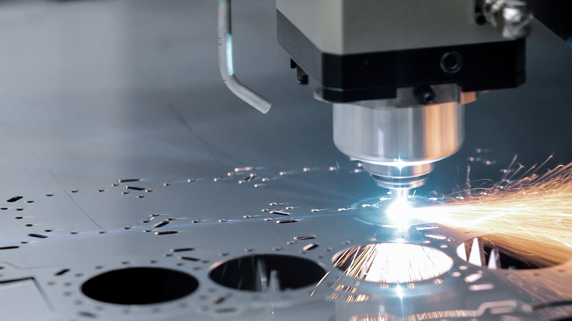 close-up of a CNC laser-cutting head slicing through a flat metal sheet, sending a cascade of bright orange sparks as the beam etches precise circular cutouts on the smooth steel surface
